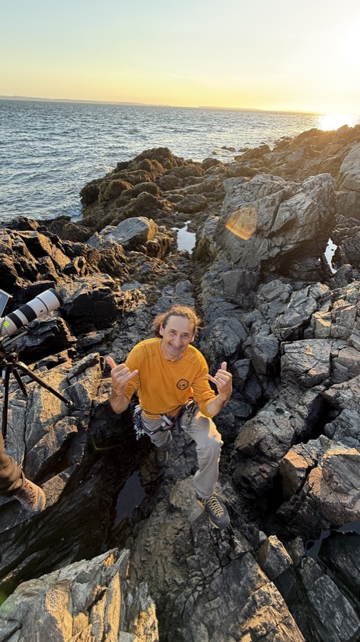 Andrea smiling on the Acadia coastline with cameras behind him.